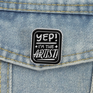 Detail of black enamel pin with Yep I'm the Artist text on denim jacket pocket