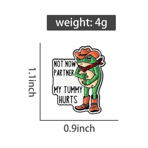 Dettaglio della spilla smaltata a rana con testo Not Now Partner My Tummy Hurts e misure