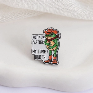 Detail view of enamel frog pin featuring cowboy hat and Not Now Partner My Tummy Hurts text