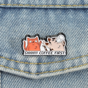 Cartoon cats enamel pin with 'SHHH!!! COFFEE FIRST' text on a denim jacket pocket