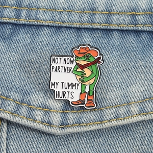 Creative cowboy frog enamel pin with Not Now Partner text on blue denim background