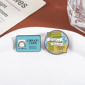 Library card and book-themed enamel pins set on a white ceramic plate and newspaper