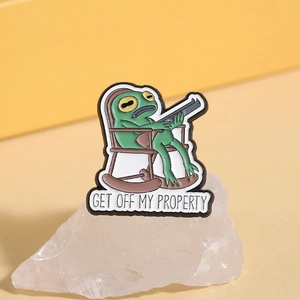 Detail der Emaille-Anstecknadel mit Frosch auf Schaukelstuhl und GET OFF MY PROPERTY Slogan