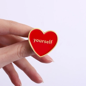 Detail showing hand holding heart enamel pin with red finish and gold yourself text
