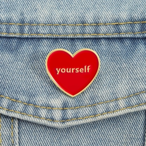 Red heart enamel brooch with 'yourself' text pinned on a blue denim jacket pocket