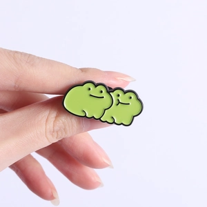 Close-up of cute green cartoon frog enamel pin held by hand for scale decoration