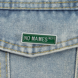 Dettaglio di una spilla smaltata verde con testo NO MAMES WEY su una giacca di jeans