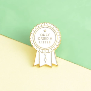 Macro view of white enamel pin with gold medal design and ONLY CRIED A LITTLE text