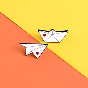 Close-up of enamel paper boat and plane pins with glitter detail and red heart accents