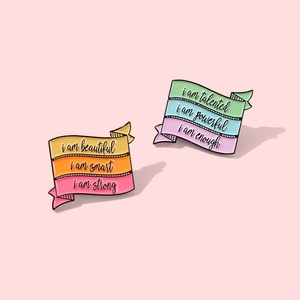 Rainbow enamel lapel pins with motivational quotes on a soft pink background