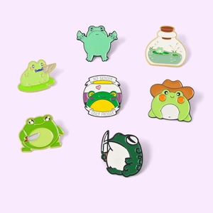 Macro detail of 7 cartoon frog enamel pins featuring green animals and slogan badges