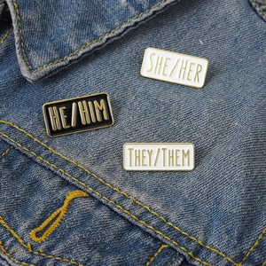 Close-up of pronoun enamel pins SHE HER HE HIM THEY THEM on blue denim jacket fabric