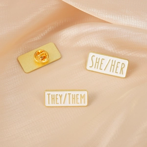 Elegant gold-plated enamel pronoun pins including She/Her and They/Them set on silk