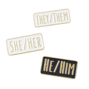Drei Emaille-Pins mit Pronomen und Goldrand mit der Aufschrift THEY, SHE und HE