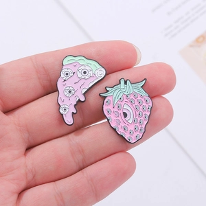 Hand holding eccentric pink pizza and strawberry enamel pins with surreal eyeball designs