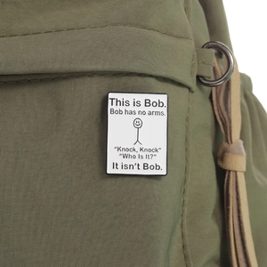 White square enamel pin featuring 'This is Bob' funny stick figure on a green canvas bag