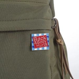 Square enamel pin featuring Back & Body Hurts text pinned on a green fabric bag