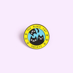 Round enamel pin featuring The Black Cat Society logo with cartoon cats on purple background