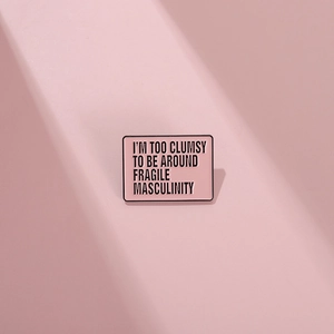 Pink rectangular enamel brooch with black text about fragile masculinity in minimalist style