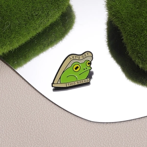 Cartoon frog bread enamel pin with 'Let's Get This Bread' slogan on a mossy background