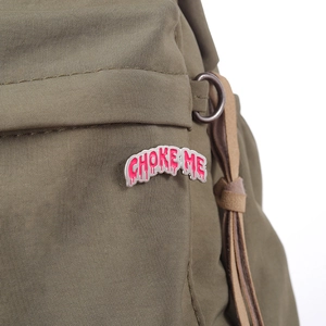 Pink enamel pin with 'CHOKE ME' text on olive green canvas background, silver metal finish