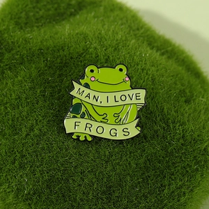 Cartoon green enamel frog pin with Man I Love Frogs banner on a mossy green surface