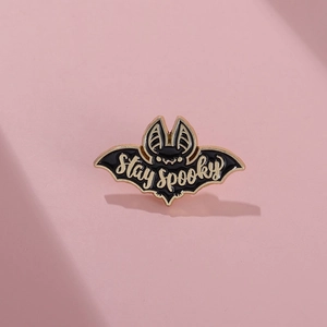 Pin's émaillé chauve-souris avec texte Stay Spooky et bordure dorée sur fond rose pastel