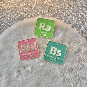 Three chemical element enamel pins with colorful alphabet designs on white plush background