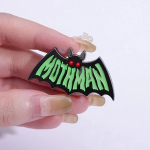 Close-up of black metal Mothman enamel pin with glowing green text and red eyes