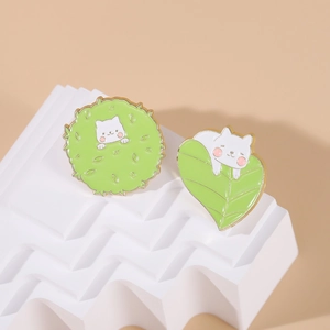 Detailed view of two cute white cat enamel pins on green leaves and bush ornaments