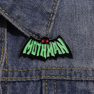 Enamel Mothman bat brooch with green lettering pinned on a blue denim jacket pocket