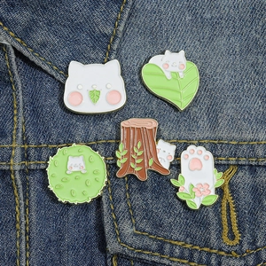 Set of 5 cartoon cat and leaf enamel pins displayed on blue denim fabric background