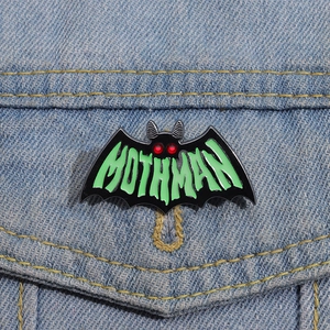 Glow-in-the-dark Mothman bat enamel pin with green lettering on denim fabric background