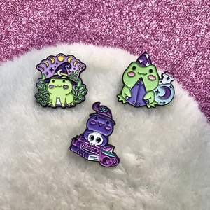 Three cute wizard frogs enamel pins with magic spell book on a pink glittery background