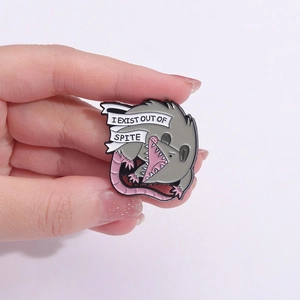 Cartoon opossum enamel pin with I EXIST OUT OF SPITE banner held in hand for scale