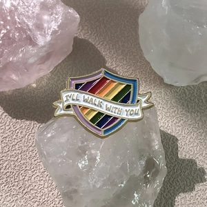 Close-up of rainbow shield enamel pin with I'll Walk With You text on crystal background