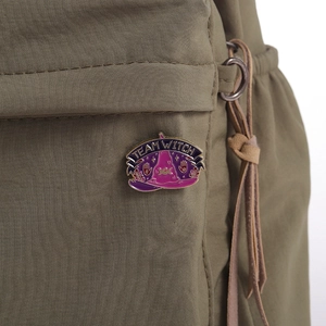 Detail view of a purple Team Witch enamel pin on a green nylon backpack strap