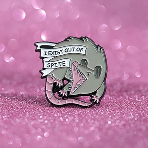 Macro shot of an angry opossum enamel pin with 'I EXIST OUT OF SPITE' text on pink glitter