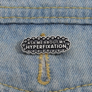 Detailed view of an oval black enamel pin on denim with Ask Me About My Hyperfixation text