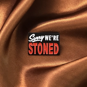 Macro detail of rectangle enamel pin with Sorry WE'RE STONED text on brown satin fabric