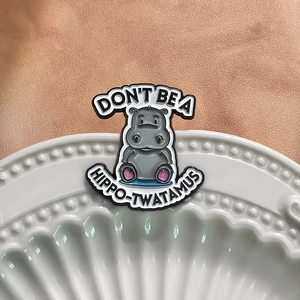 Funny grey cartoon hippo enamel pin on a white plate with Hippo-Twatamus text slogan
