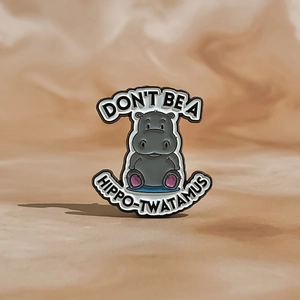 Cartoon hippo enamel pin with Don't Be A Hippo-Twatamus slogan on textured background
