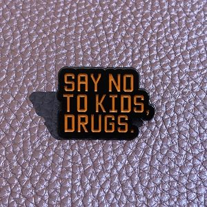 Black enamel brooch with orange Say No To Kids, Drugs text on purple leather background