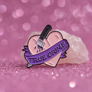 Dettaglio della spilla smaltata a pugnale True Crime con cuore su sfondo rosa glitter