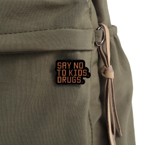 Close-up of satirical 'SAY NO TO KIDS, DRUGS' enamel pin on an olive green nylon backpack