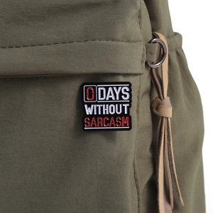 Detail of 0 Days Without Sarcasm enamel pin on army green fabric with leather tassel