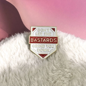 Detail of shield enamel pin with Don't Let The Bastards Grind You Down lettering