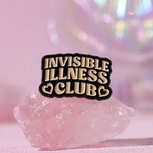 Makroaufnahme der Emaille-Anstecknadel Invisible Illness Club auf rosa Kristall