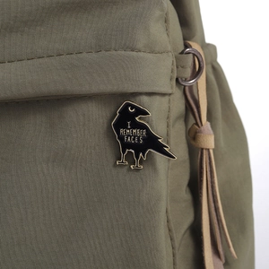 Detail of a black and gold raven enamel pin with I REMEMBER FACES text on a fabric bag