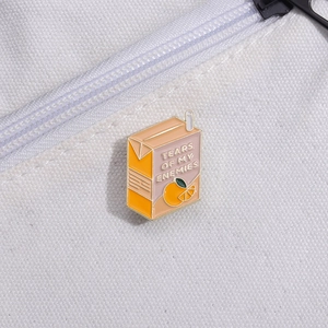 Detail of cartoon orange milk box enamel pin with 'Tears of My Enemies' text on canvas